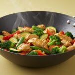 Colorful chicken stir fry with broccoli, zucchini, red bell pepper, and snap peas in a black wok on a yellow background.